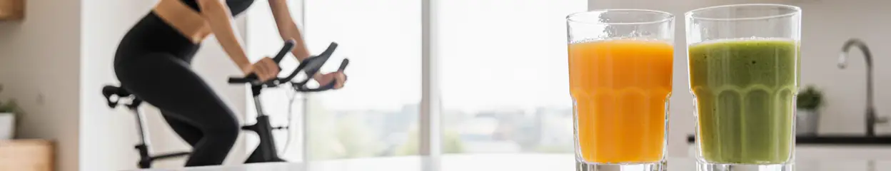 Vrouw sport op hometrainer naast twee glazen met groente- en fruitsap.